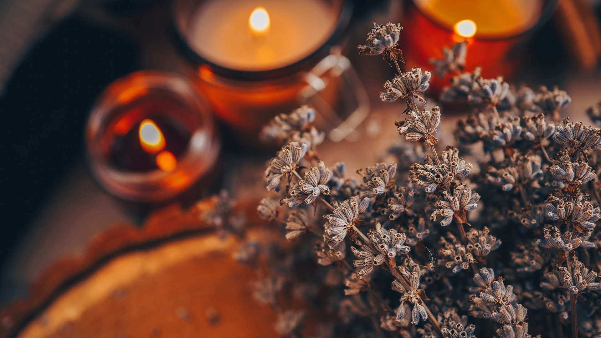 Close-up of dried lavender flowers with three lit candles in amber holders softly blurred in the background.